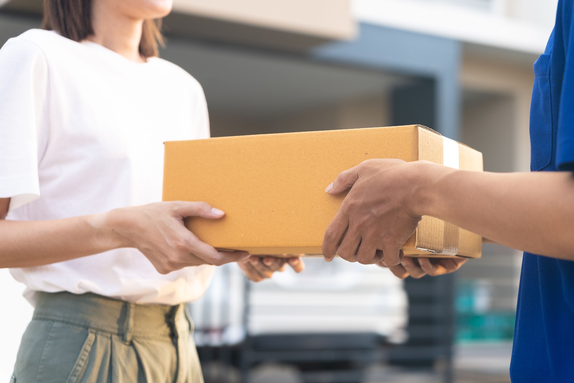 Close up delivery man hands wearing blue uniform send a cardboard box to customer in front of the customer's house with sunset flare background.